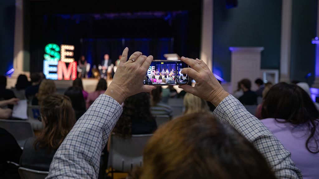 A man holding up a smartphone taking a picture of the stage at the SEEM Conference.