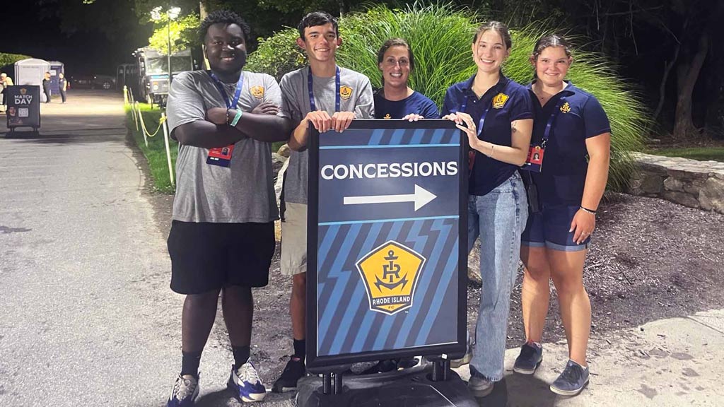 JWU students and alums Soh Kone, Jack Sacco, Allison Pangakis, Rain Keefe and Michelle Charron post around a concessions sight at a 2024 RI Football Club match